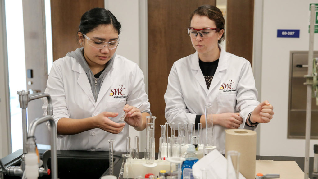 Two college students wear lab coats and handle lab equipment