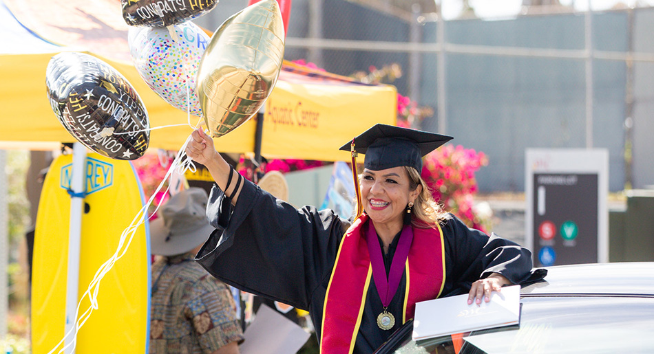 A woman wearing a graduation gown excitedly waves with balloons